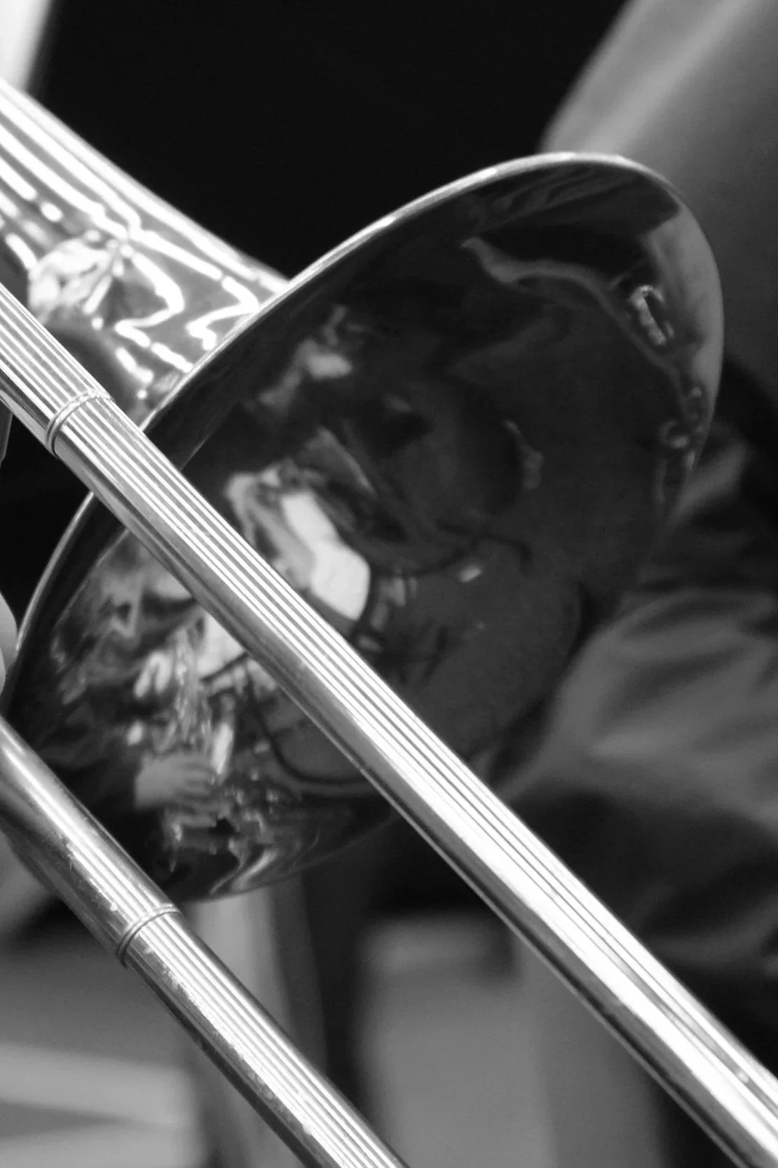 Close-up of a brass instrument, possibly a trumpet or trombone, with a shiny metallic surface and in focus. Another tube, likely part of the same instrument, is visible crossing in front of it.