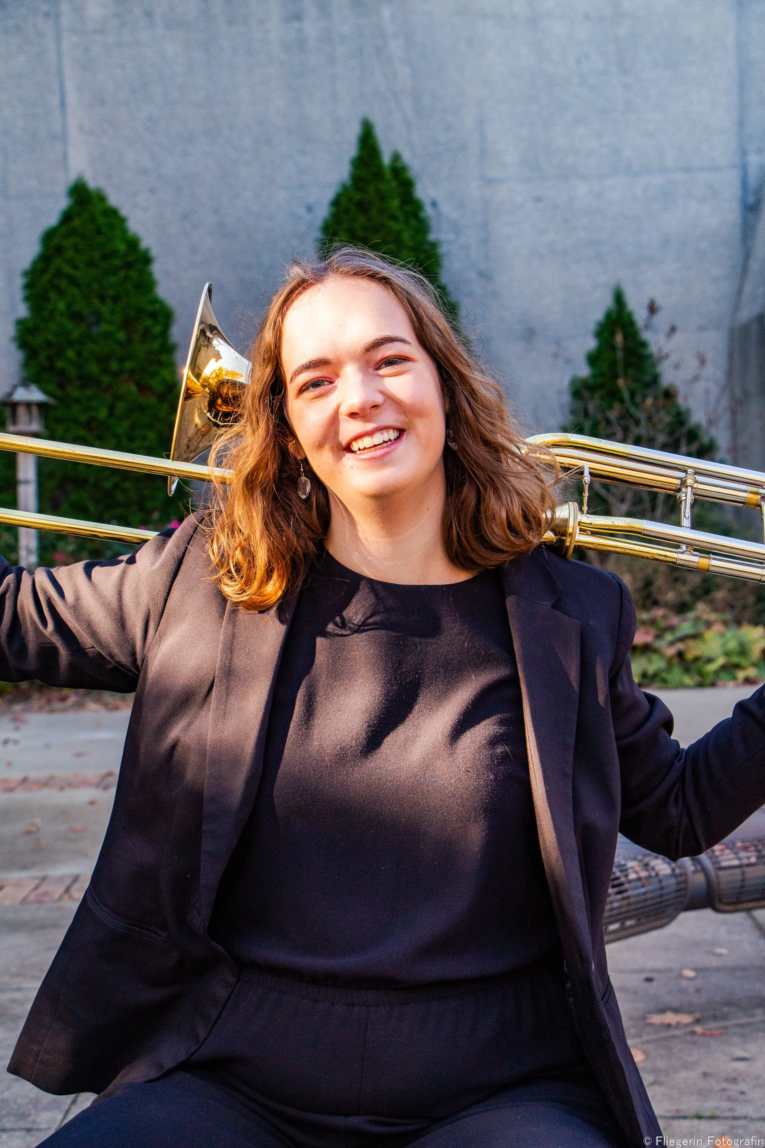 A young woman with long, wavy brown hair and earrings smiling playfully outdoors, holding a brass trombone over her shoulder, with a background of tall shrubs and a grey wall.