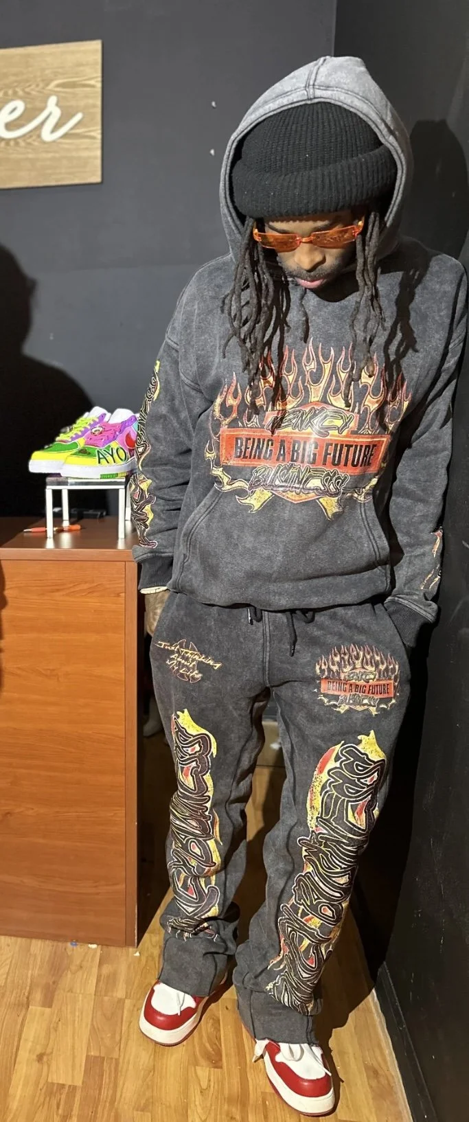 A man dressed in a black hoodie, black beanie, and red sunglasses, standing indoors with his head bowed. He is wearing a dark grey graphic hoodie and matching sweatpants with colorful flame and graffiti-style designs. There is a pair of vibrant, multicolored sneakers on display on a small table in the background.