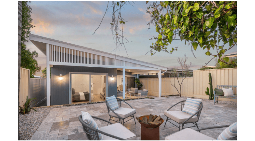 Outdoor patio with seating area, fire pit, and view of a modern house with sliding glass doors and a covered porch, surrounded by a wood fence and trees.
