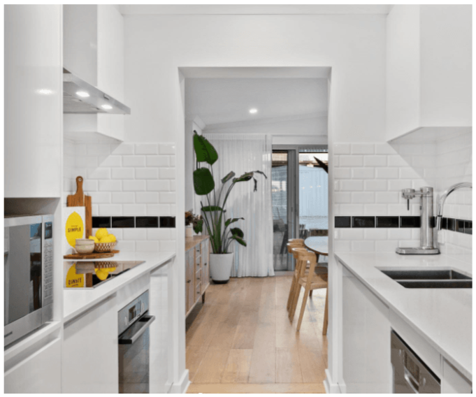 Modern kitchen with white cabinets, black and white tiled backsplash, wooden floor, and a large potted plant near the door leading to a patio.