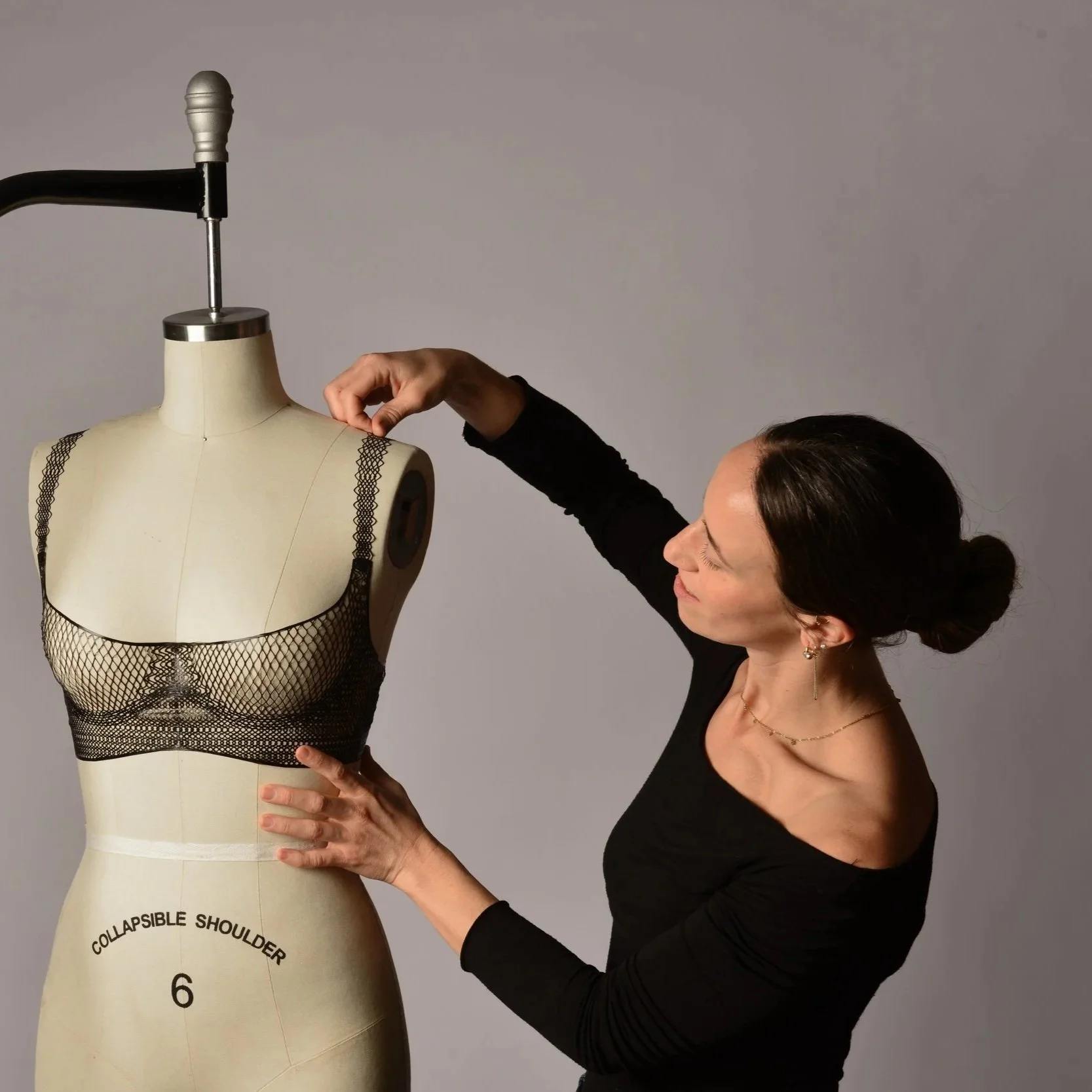 A woman adjusting a black lace dress form mannequin on a white stand against a plain gray background.