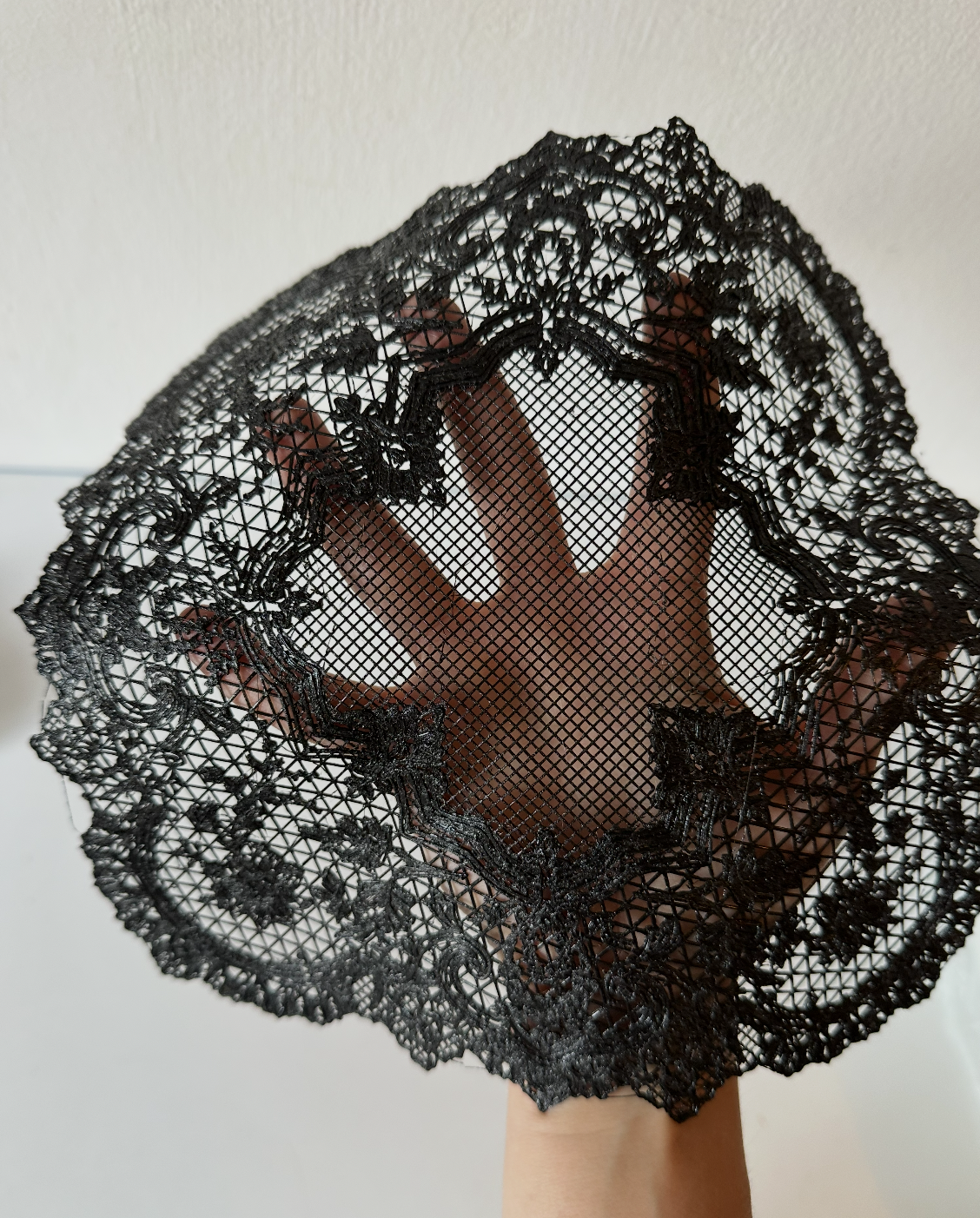 A hand holding up a black lace umbrella with intricate floral patterns and a scalloped edge against a plain light-colored wall.