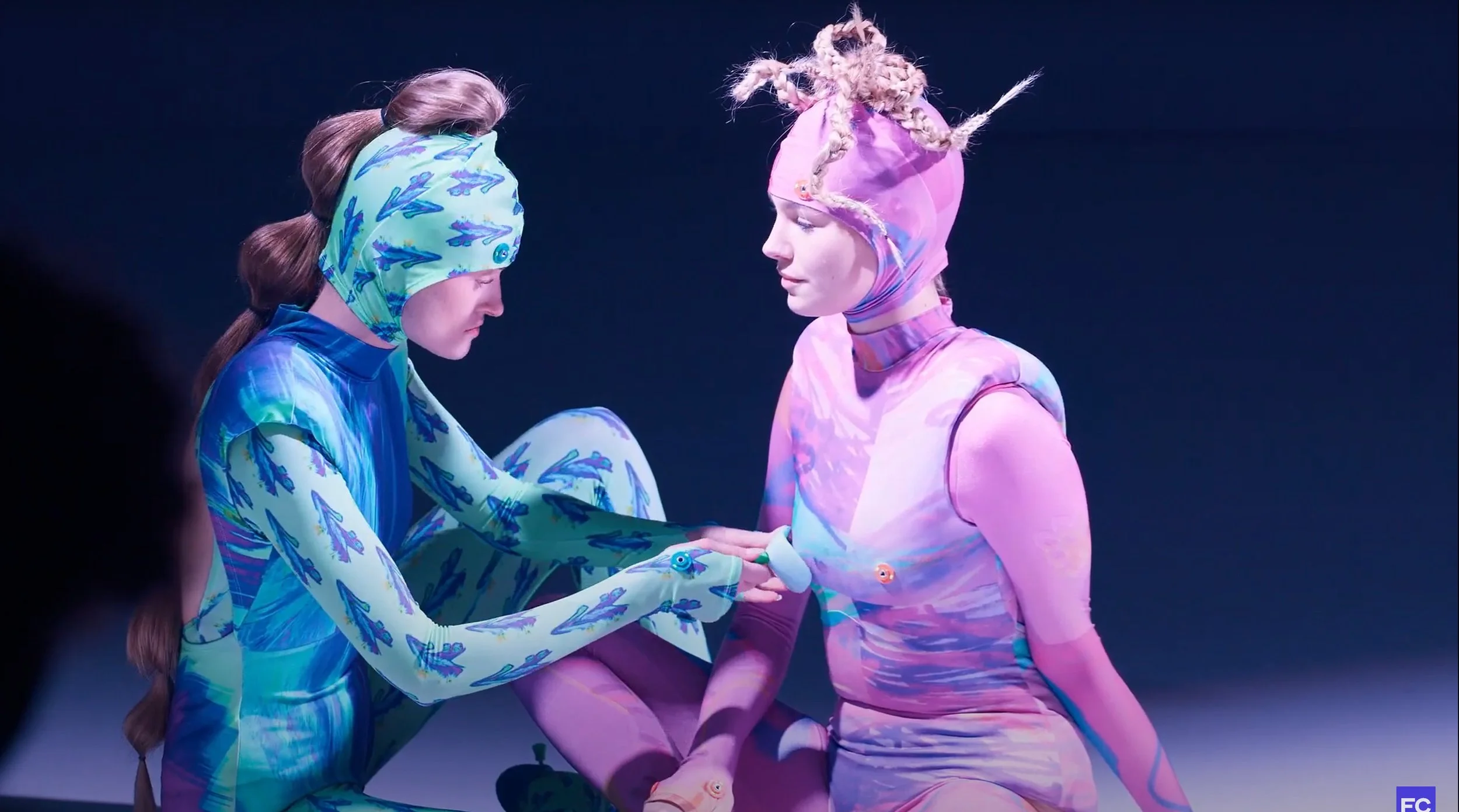 Two individuals dressed in colorful, patterned costumes with one person holding the other's hands, set against a dark background.