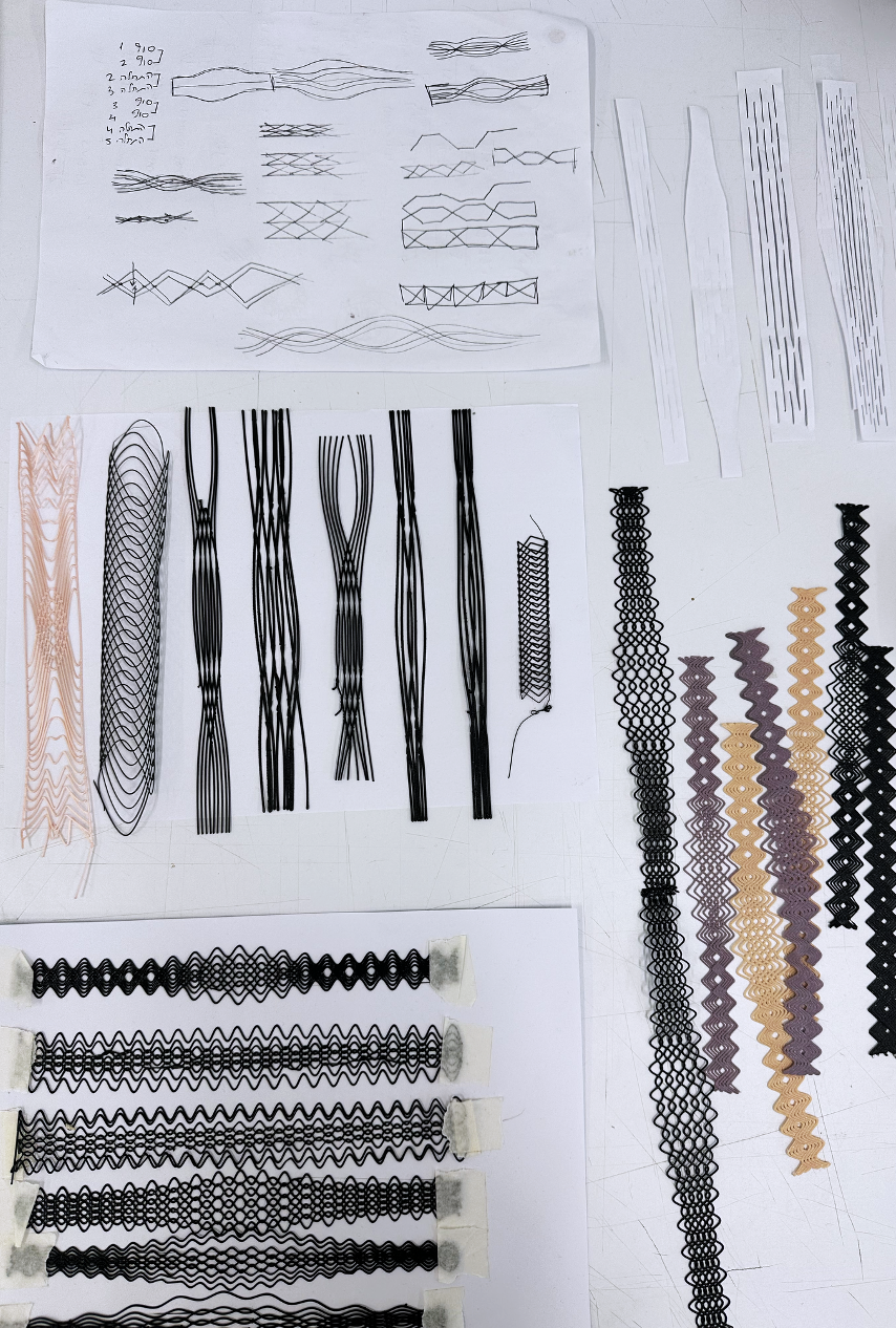 Sheet of paper with sketches and plans for weaving or lace patterns, alongside samples of intricate woven or lace strips in black, beige, and purple on a white surface.