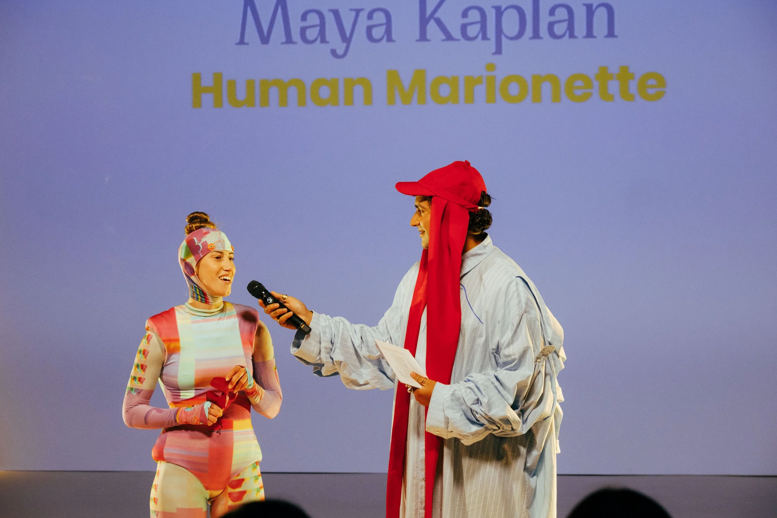 Two individuals on stage, one woman wearing colorful striped outfit and a matching hat, and one man wearing a white jacket with a red scarf and a red cap, engage in a conversation with a microphone, with a blue background displaying the name Maya Kapran and the words 'Human Marionette'.