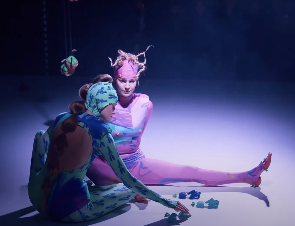 Two performers in colorful costumes sit on the floor in a dark space with a spotlight, surrounded by toys and a hanging doll. One performer has a pink and purple costume with dreadlocks and the other has a blue and green costume with a head covering.