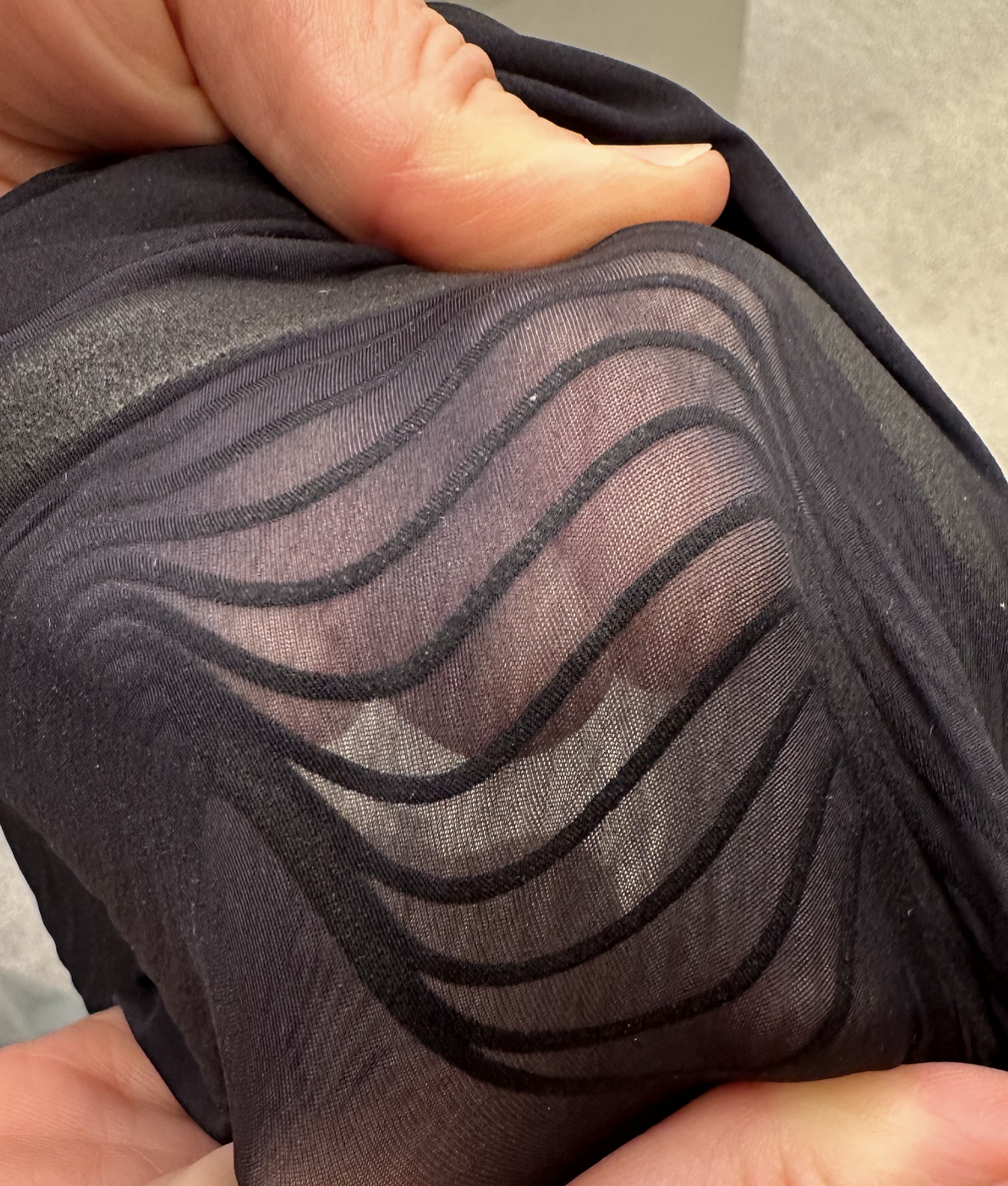 Close-up of a person's hand holding a piece of black, sheer fabric with wavy striped pattern, revealing the skin underneath.