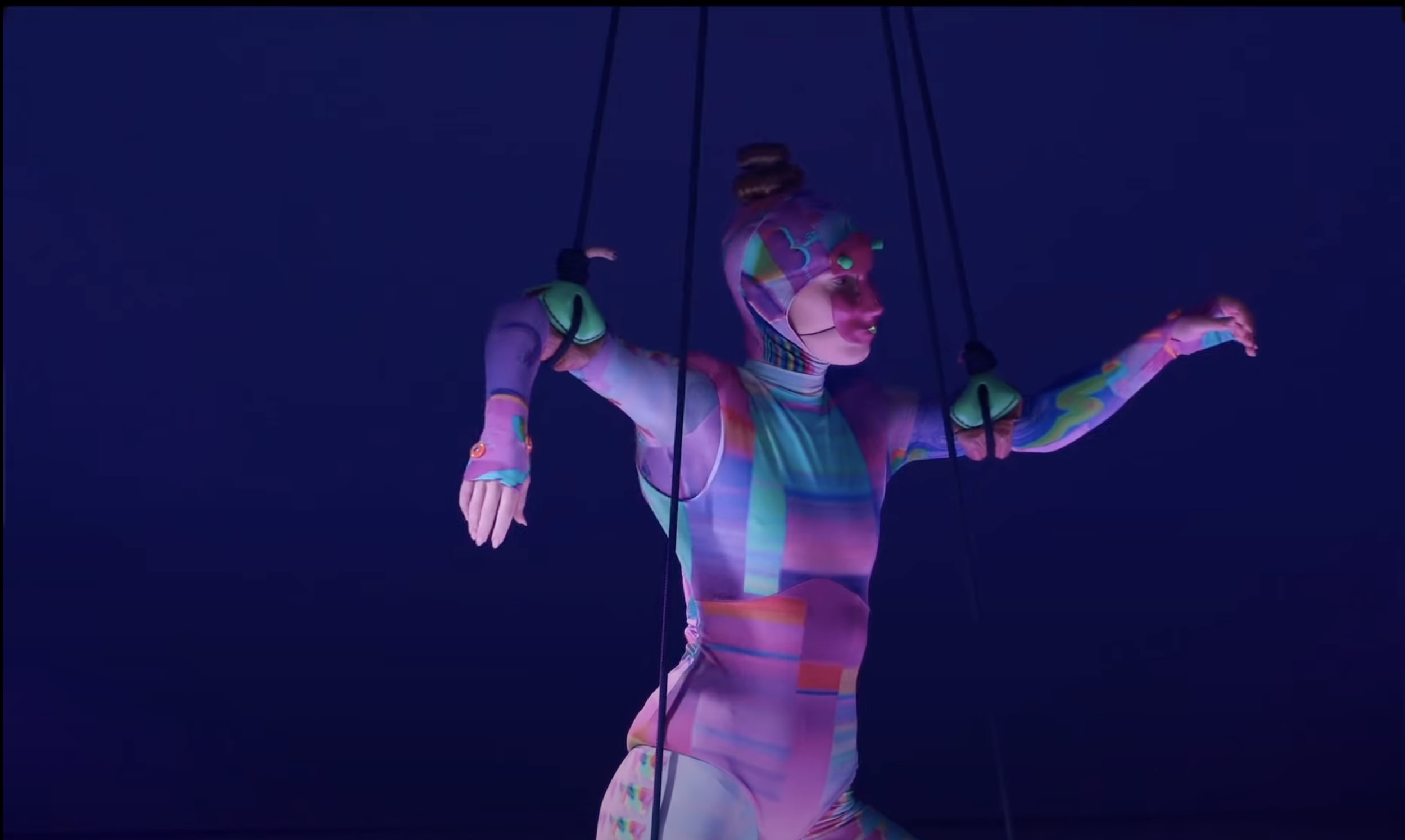 A performer on a dark stage, dressed in a colorful, rainbow-patterned bodysuit, suspended with harnesses and ropes, with their arms extended.