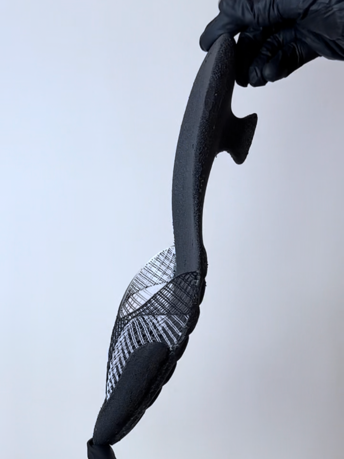 A modern black and gray skateboard with a textured grip tape surface held in midair by a hand wearing a black glove, against a plain white background.