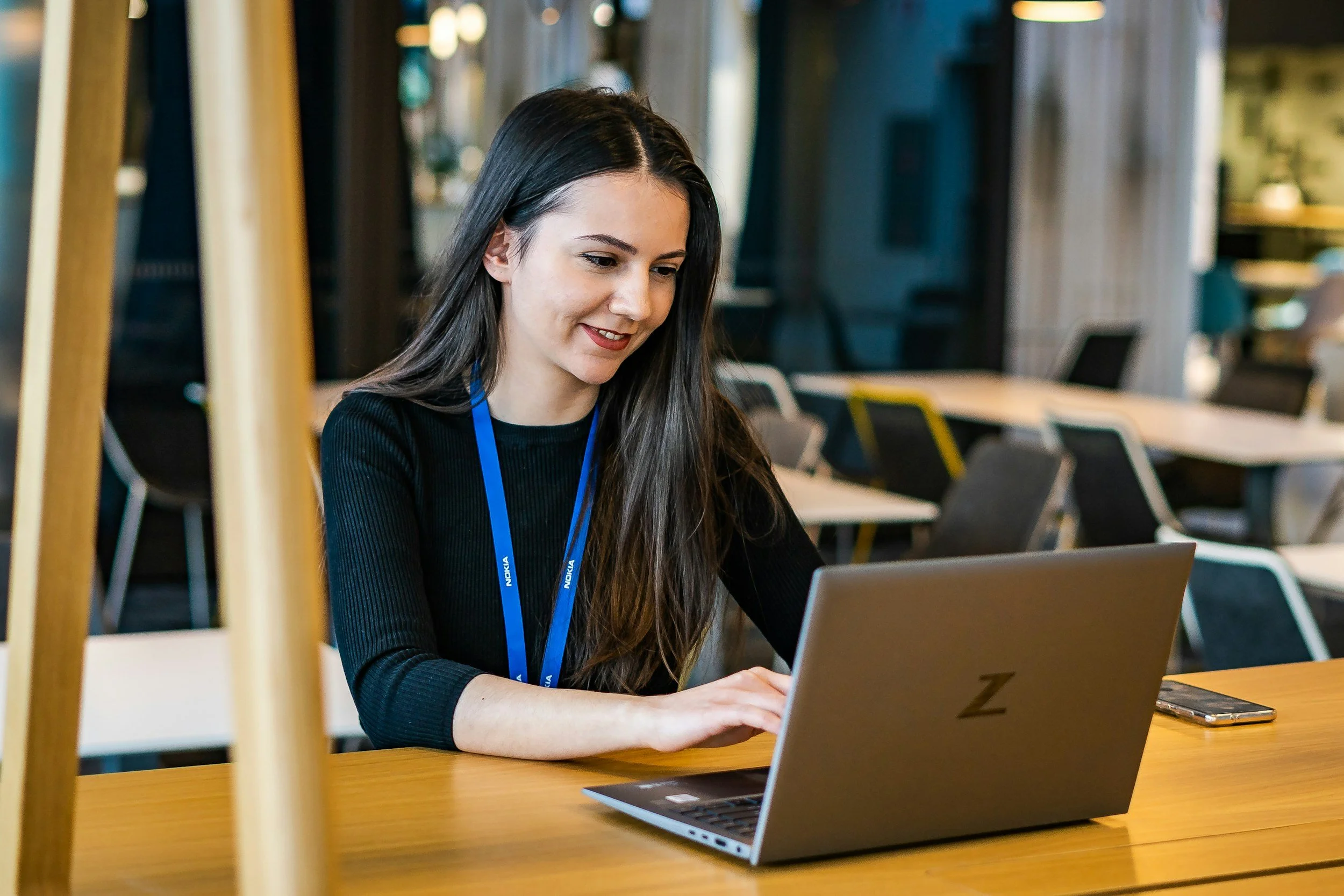 A young woman with long dark hair working on a laptop at a table in a modern, well-lit workspace or conference room. She has a smile and is focused on the screen. She is wearing a black top and a blue lanyard. The background features chairs and tables, with a blurred view of the room interior.