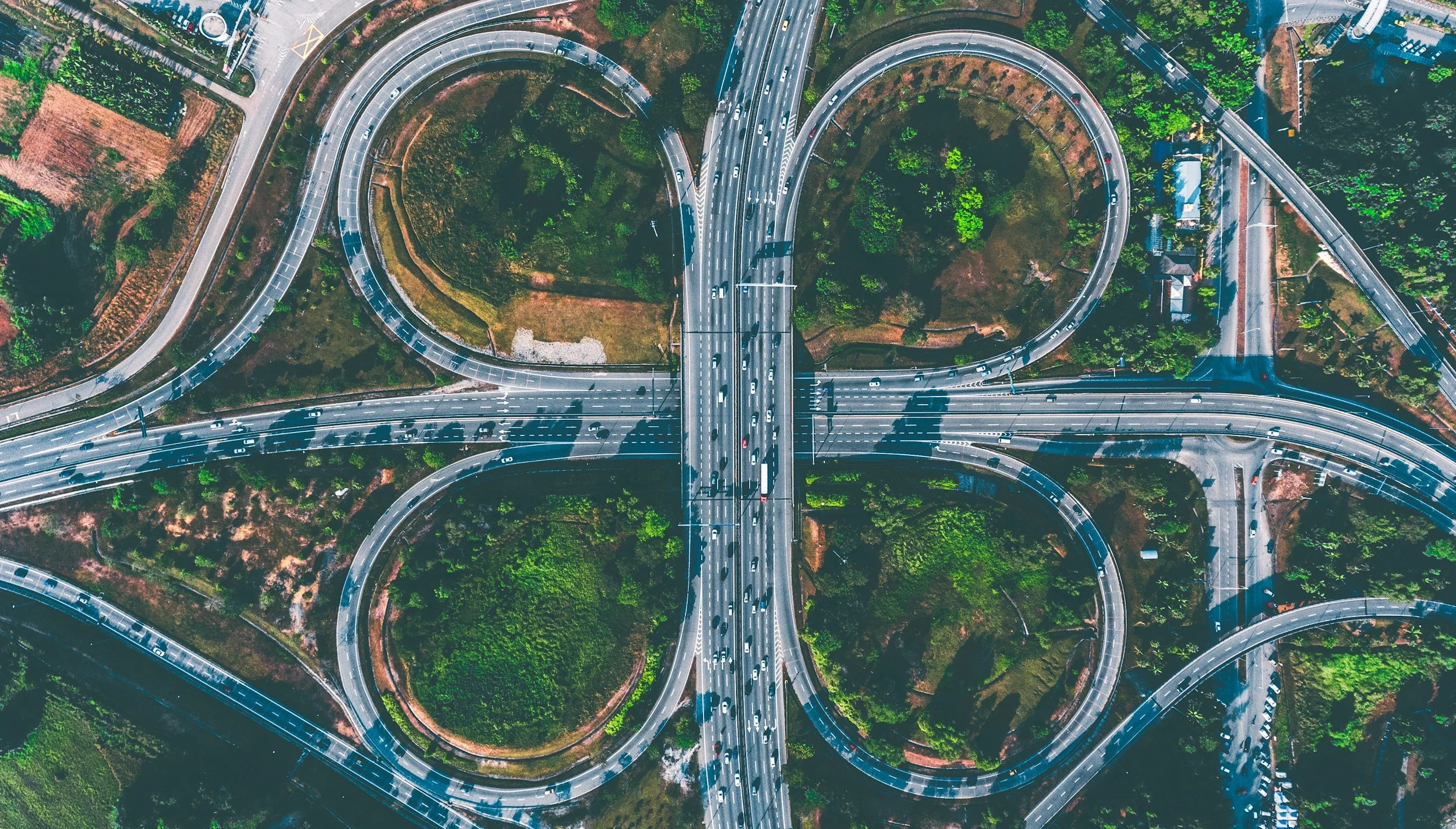 An aerial view of a complex highway interchange with multiple curved ramps and roads, surrounded by green trees and some buildings.
