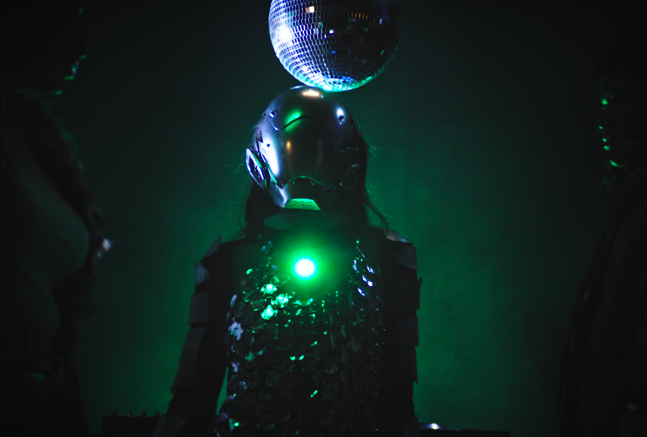 Person in futuristic costume with a robotic mask and sequined outfit, standing under a disco ball with green lighting at a dance party.