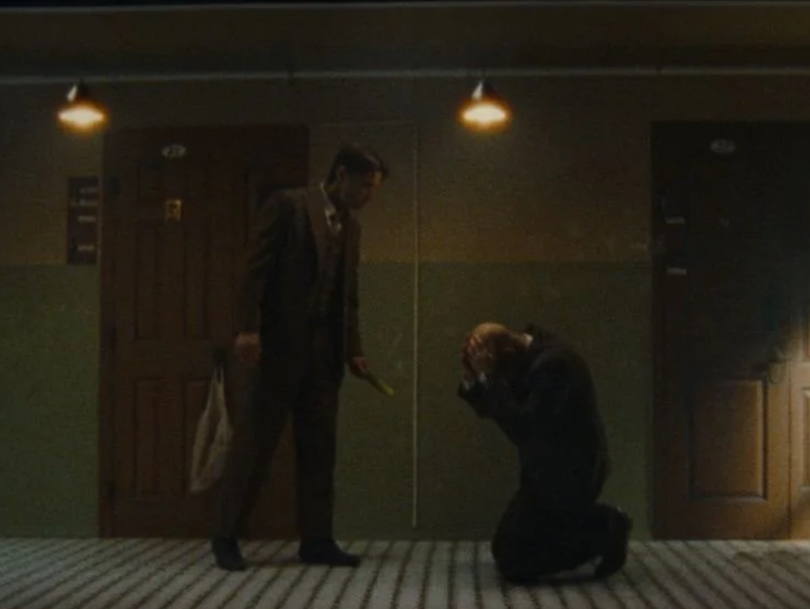 A person in a suit standing while another person is kneeling and taking a photo in a dimly lit hallway with doors and overhead lights.