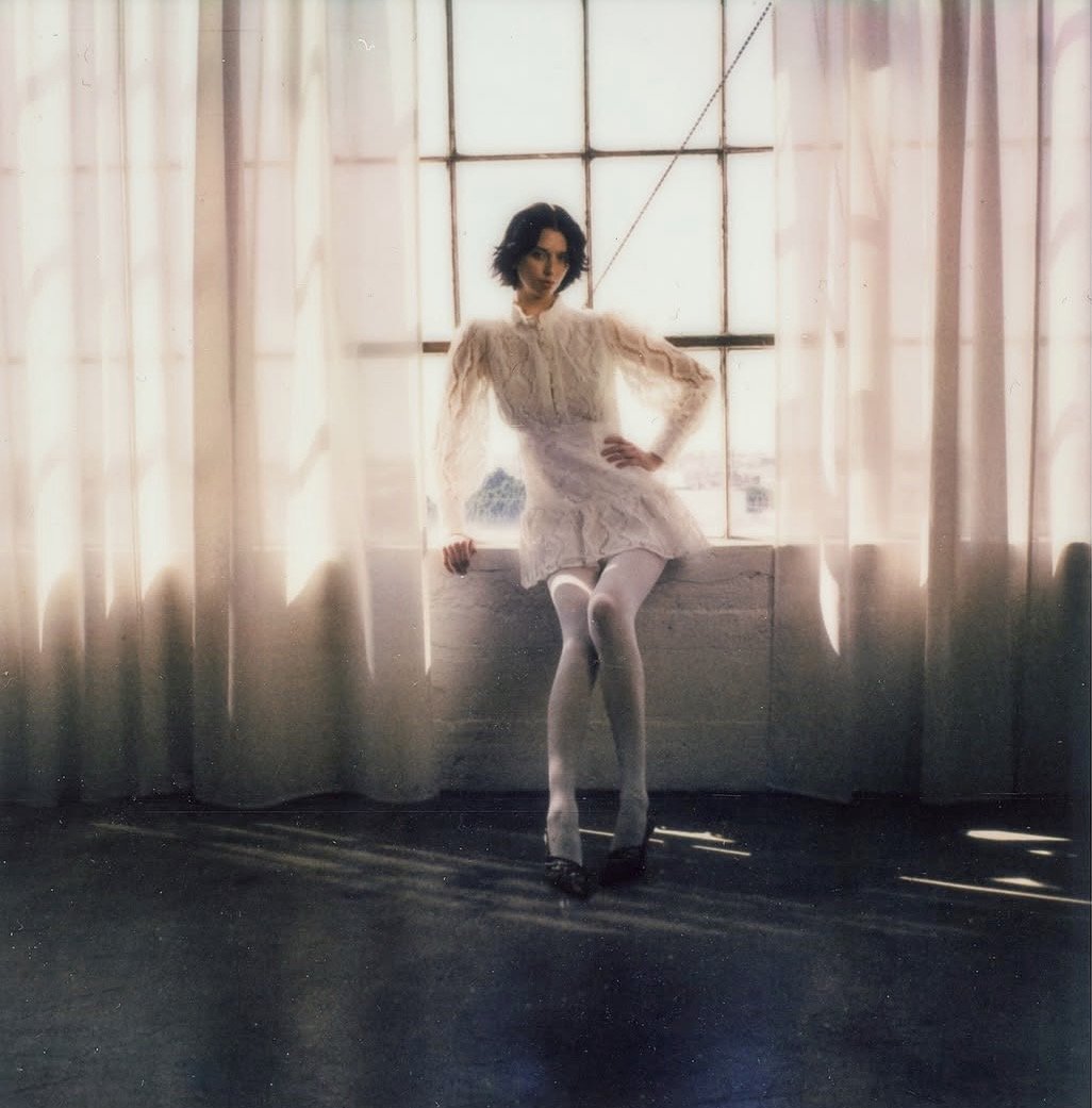 A woman with short dark hair wearing a cream-colored dress with puffed long sleeves, stockings, and high heels, standing in front of a large window with sheer curtains, bathed in soft natural light.