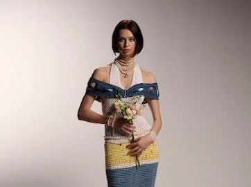 A woman wearing a multicolored striped dress holding a small bouquet of flowers against a plain gray background.