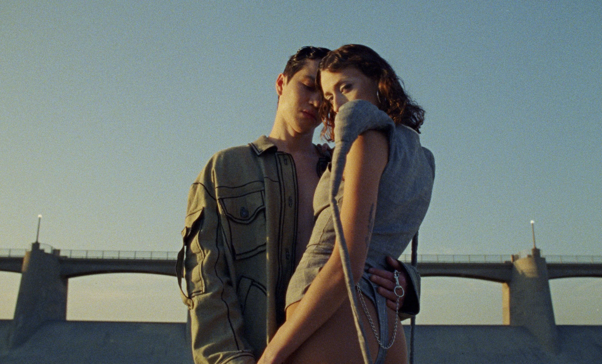 A young couple stands close together on a bridge at sunset, with the man gently resting his forehead on the woman's head.