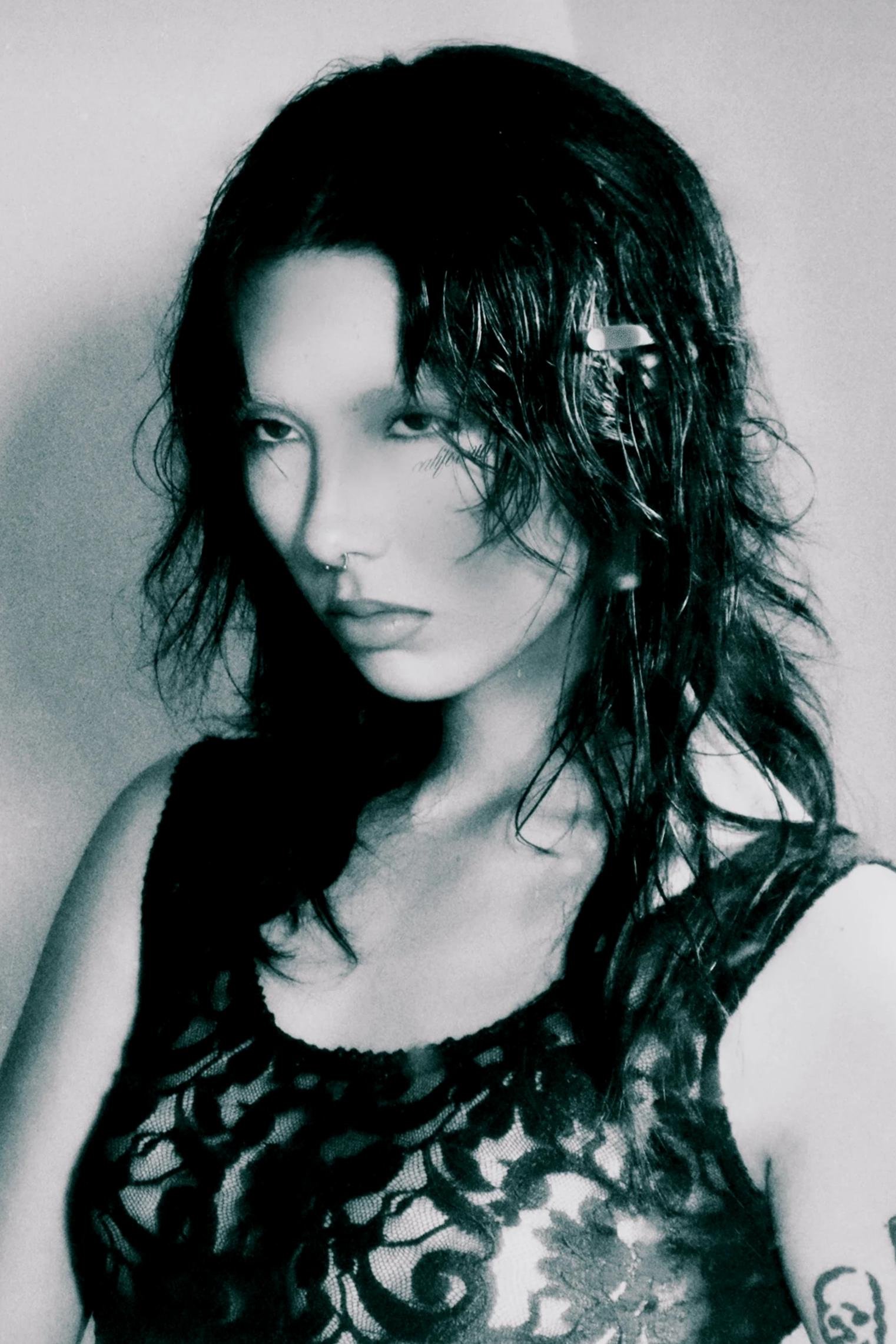 A woman with dark, wavy hair and a septum piercing is looking at the camera. She is wearing a black leather and fishnet top and has a tattoo on her arm.