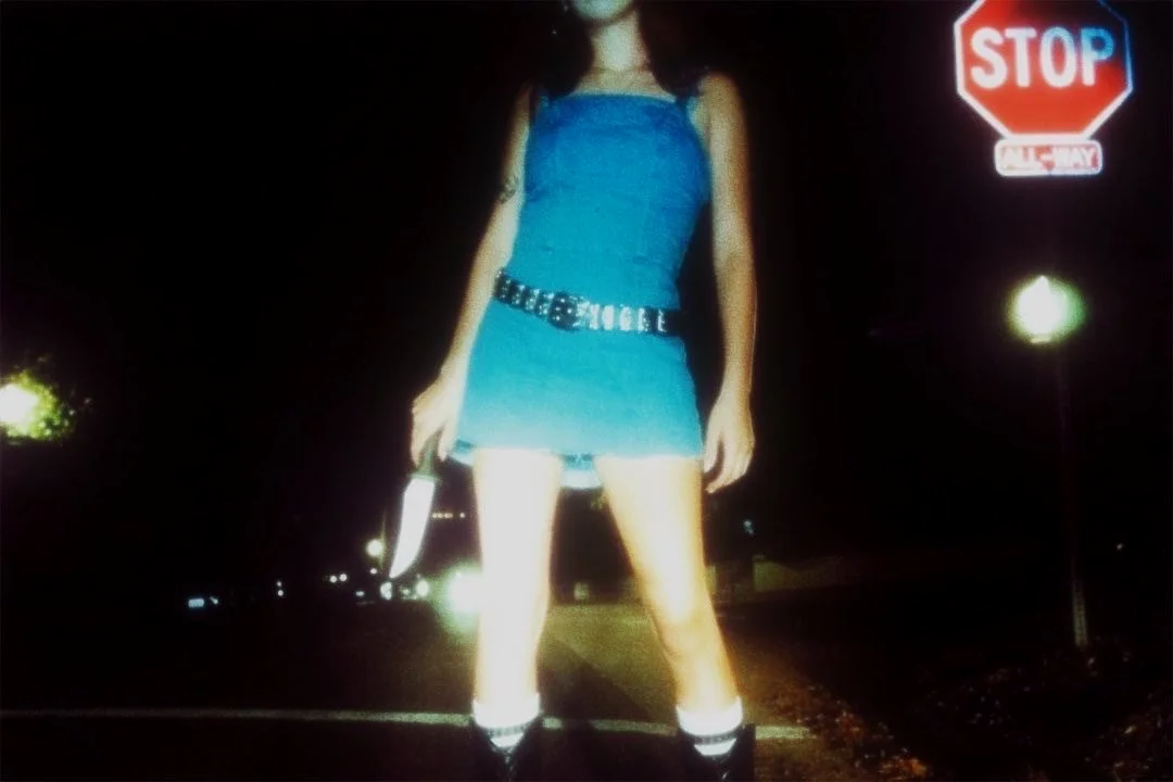 A young woman standing in the street at night, wearing a blue dress with a belt, holding a knife in her right hand, with a stop sign visible in the background.