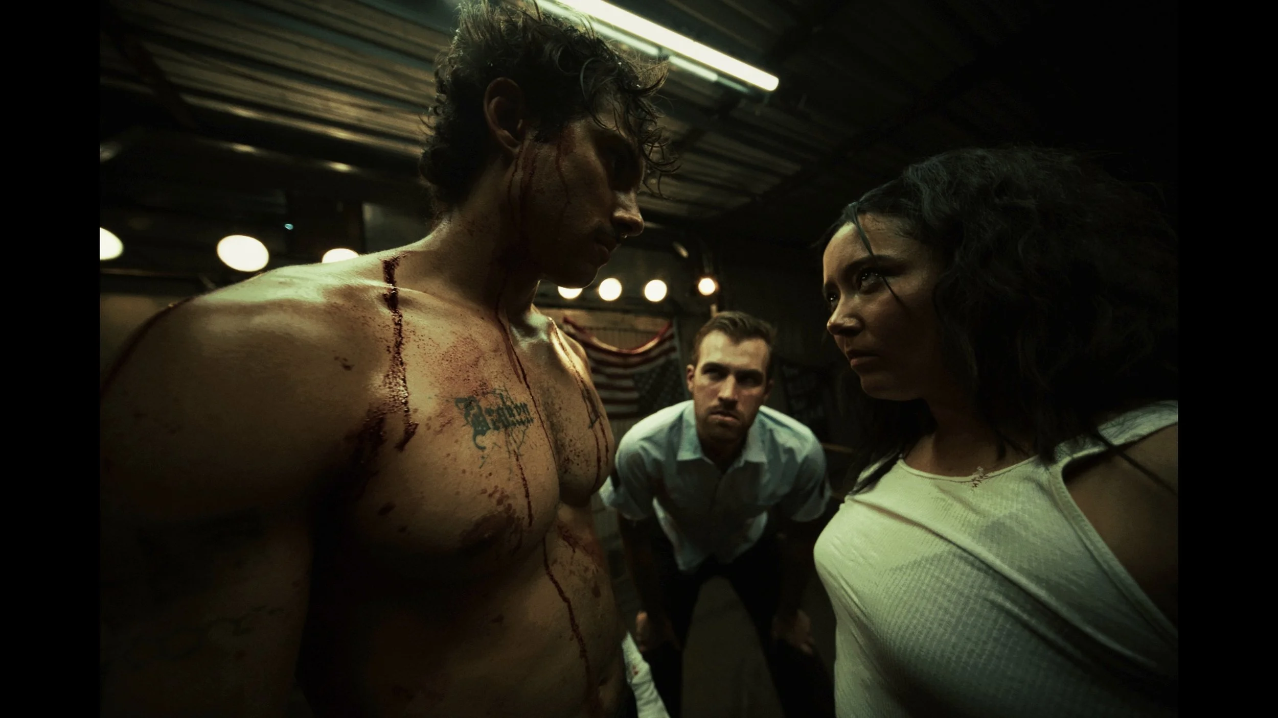 A muscular man with blood and scratches on his body, with a tattoo on his chest, looks intensely at a woman with disheveled dark hair and a white shirt, while two men in the background observe in a dimly lit, industrial setting.