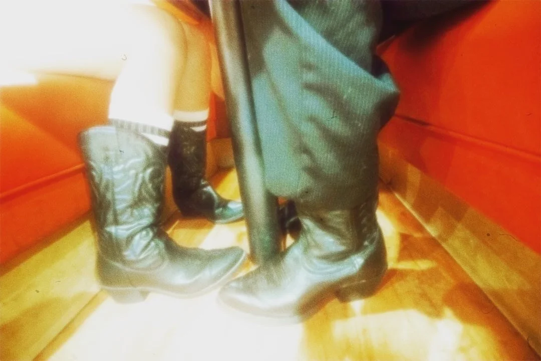 Two pairs of black cowboy boots and one pair of black leather shoes on a wooden floor between two red furniture pieces.