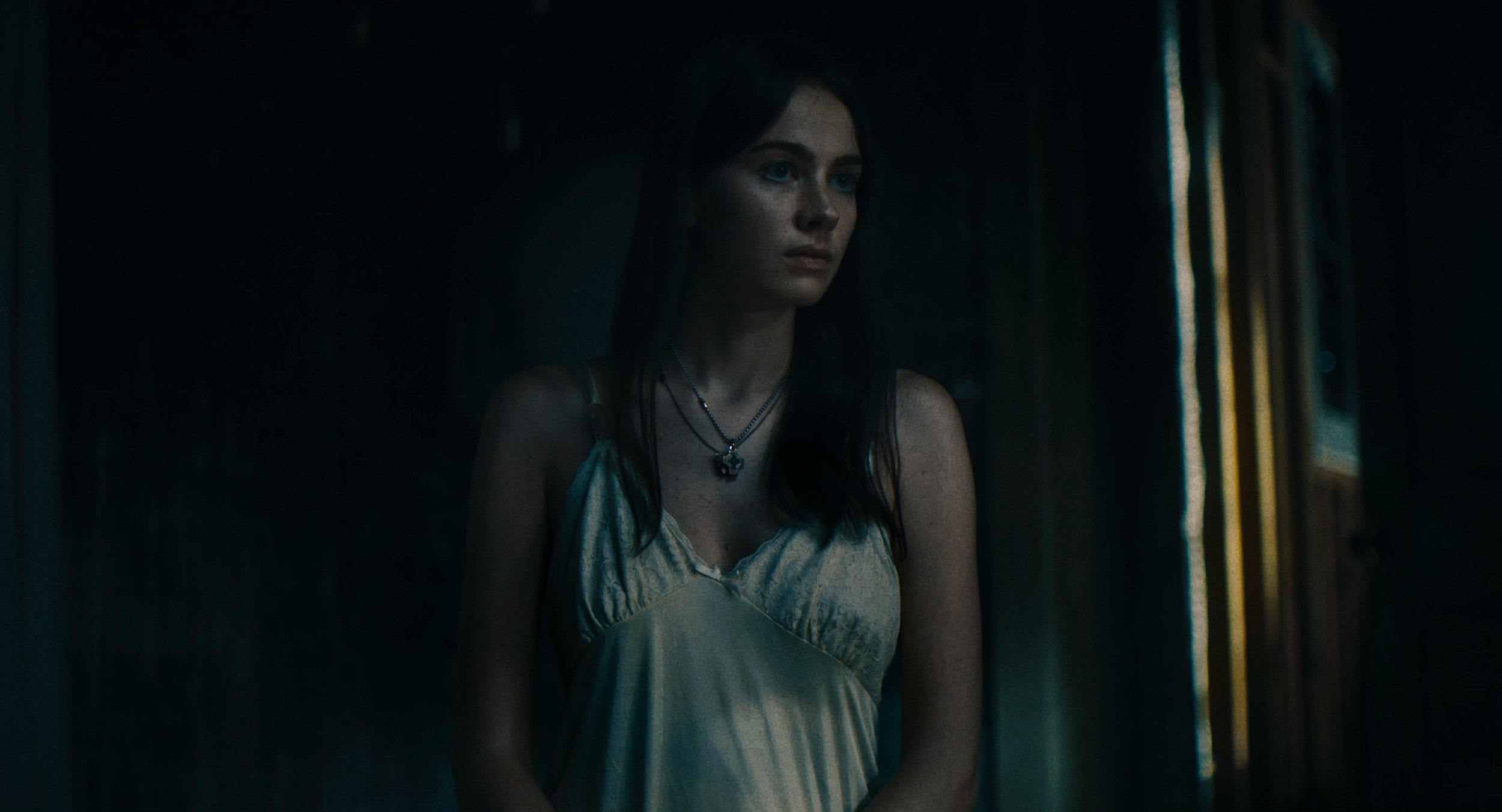 A young woman with dark hair and blue eyes wearing a white satin slip dress, standing in a dimly lit room by a window with blinds.