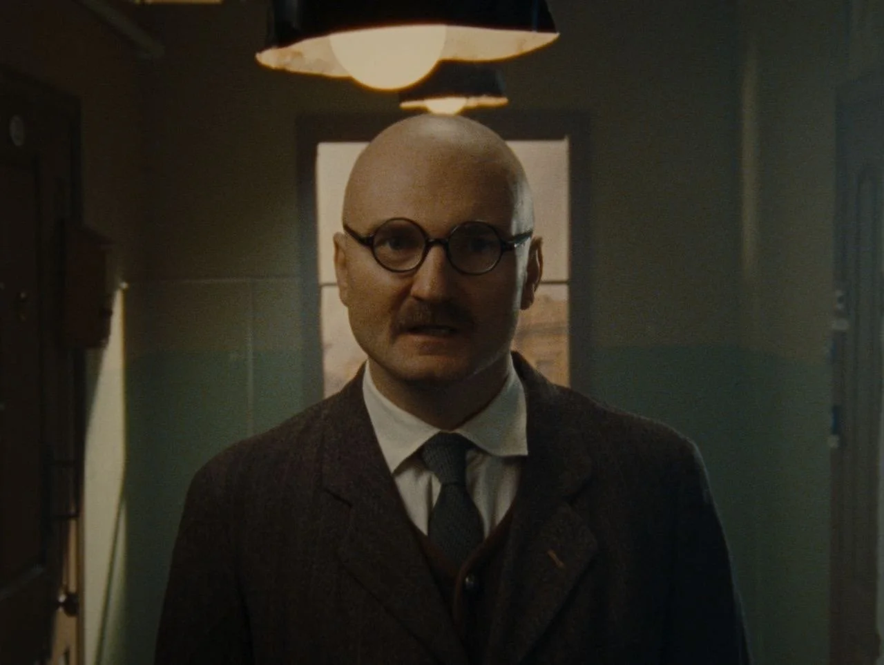 A bald man wearing round glasses, a suit, and a tie, standing in a dimly lit room with a window behind him.