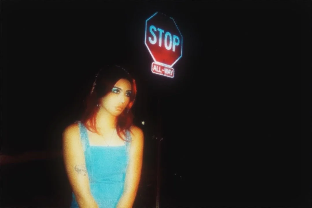 A woman with red and dark hair standing in front of a dark background with neon signs, including a red stop sign with blue letters and the word 'STOP' and a smaller sign reading 'ALL-WAY'.