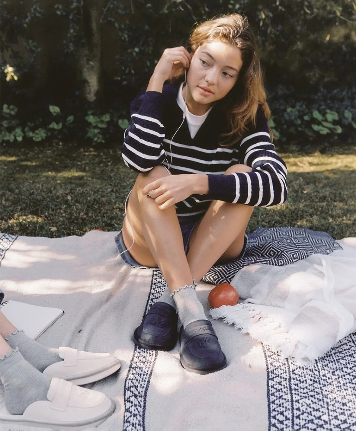 Young woman with wavy brown hair sitting outdoors on a blanket, wearing a navy and white striped sweater, light shorts, and gray socks with black slip-on shoes, listening to music with earbuds, with a red apple and white cloth nearby.