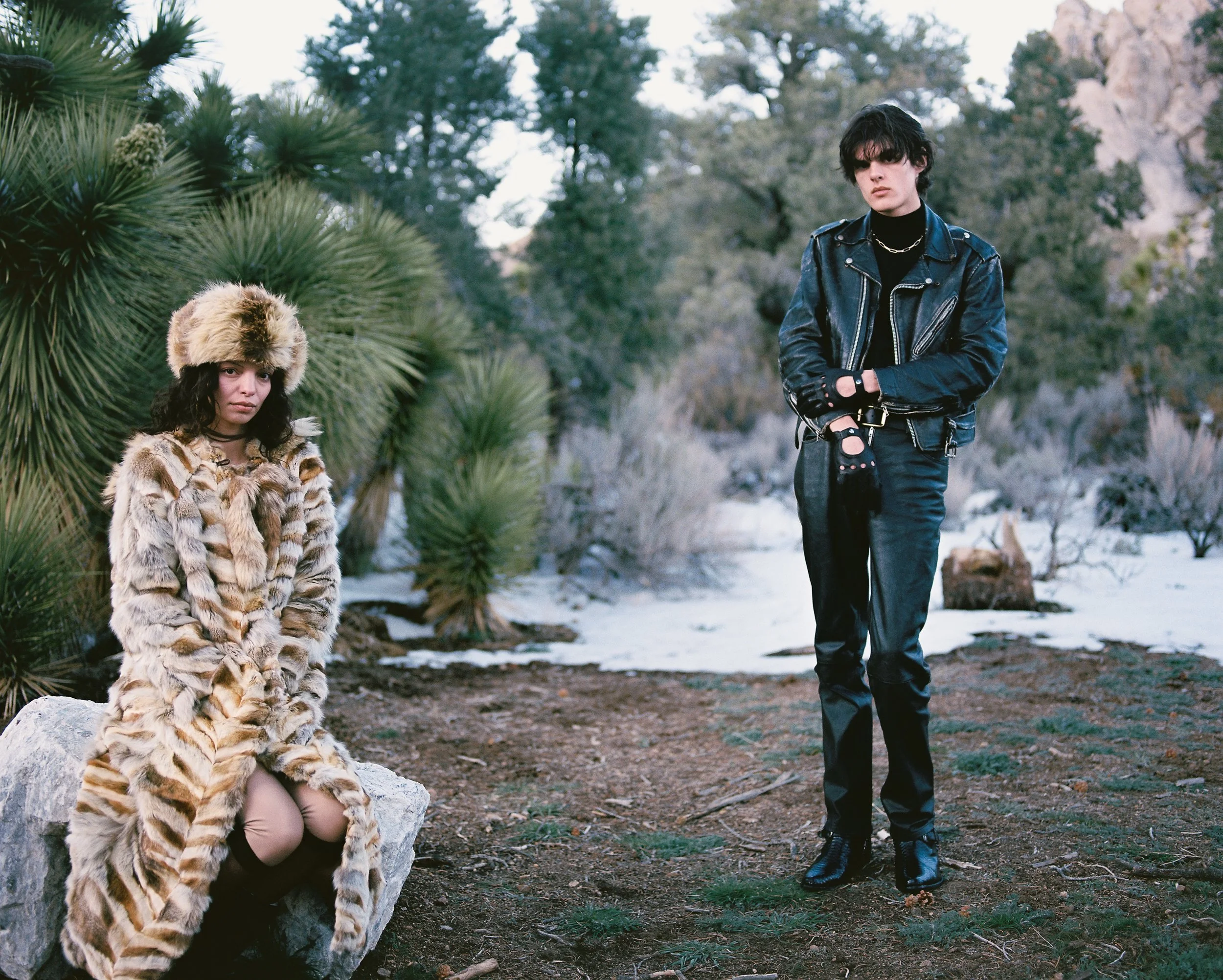 A woman wearing a fur coat and hat sitting on a rock, and a man in a leather jacket and black gloves standing outdoors in a natural setting with trees and rocks.