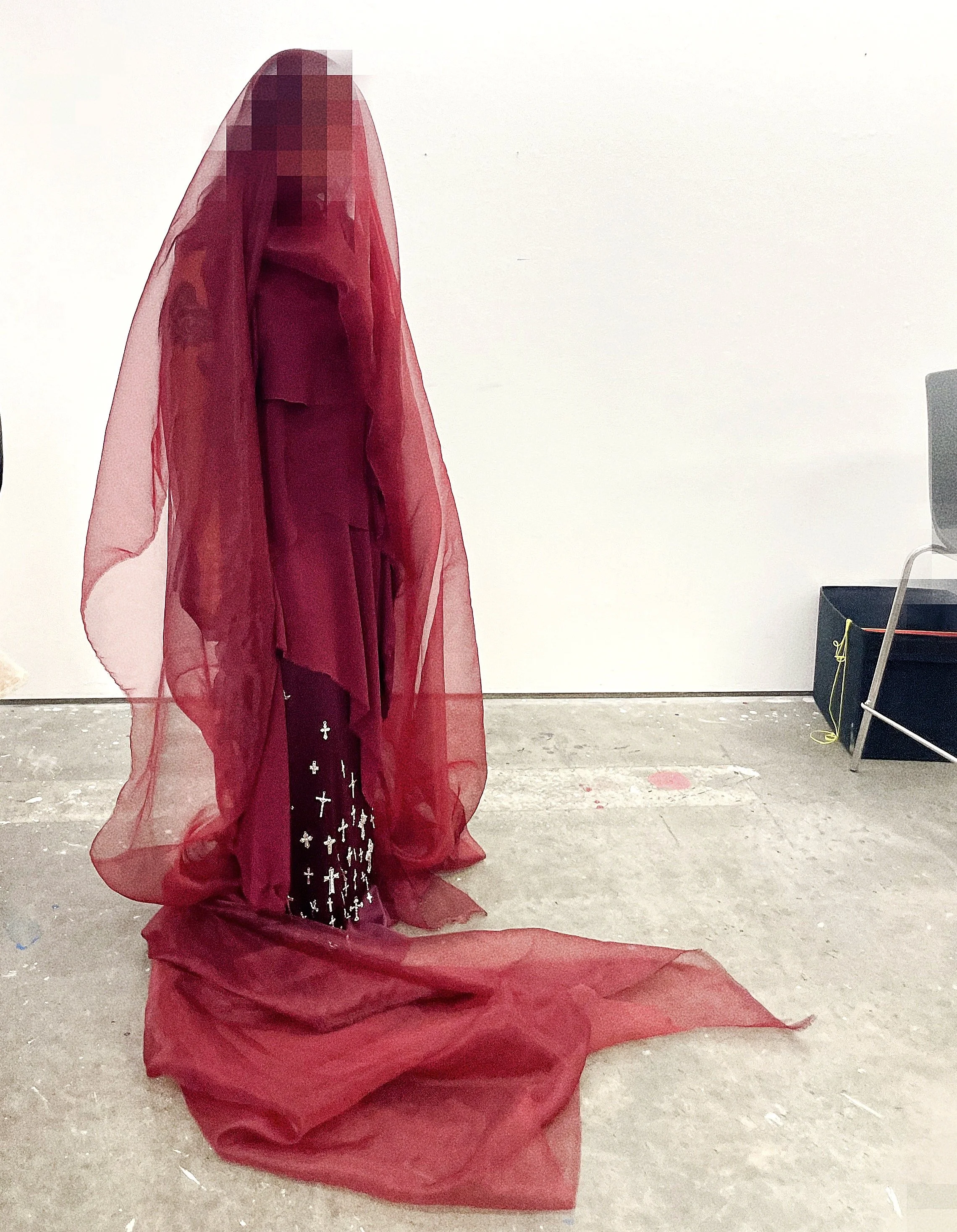 A person dressed in a long black dress with white crosses, wearing a red sheer cape and a red veil covering their face, standing in a room with a plain white wall and a chair.