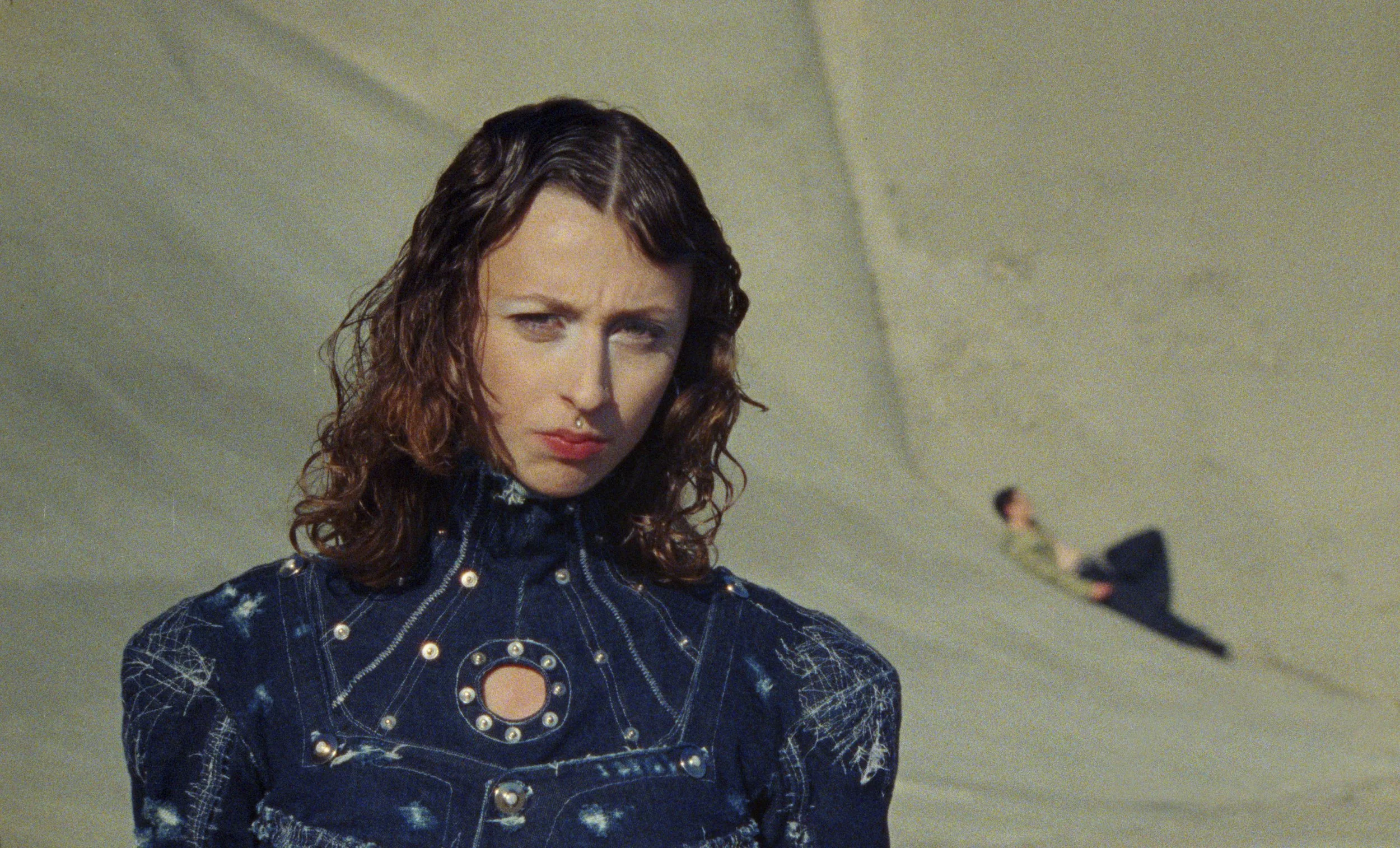A woman with curly hair and a nose piercing wearing a dark patterned denim shirt.