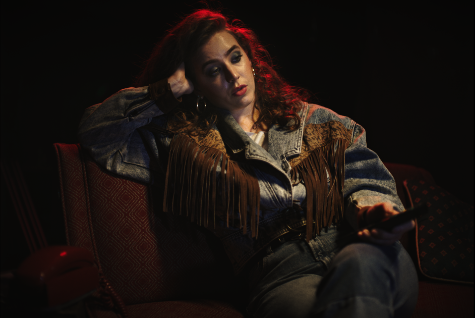 A woman with long, wavy brown hair sits on a dark red patterned couch, wearing a denim jacket with fringes and looking at her phone in a dimly lit room.