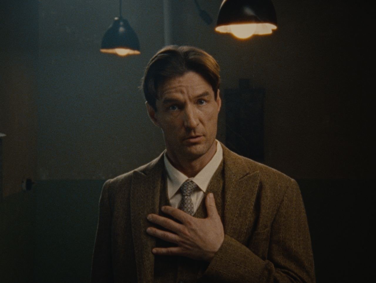A man in a brown blazer and white shirt with a tie, holding his chest, standing in a dimly lit room with hanging ceiling lights.