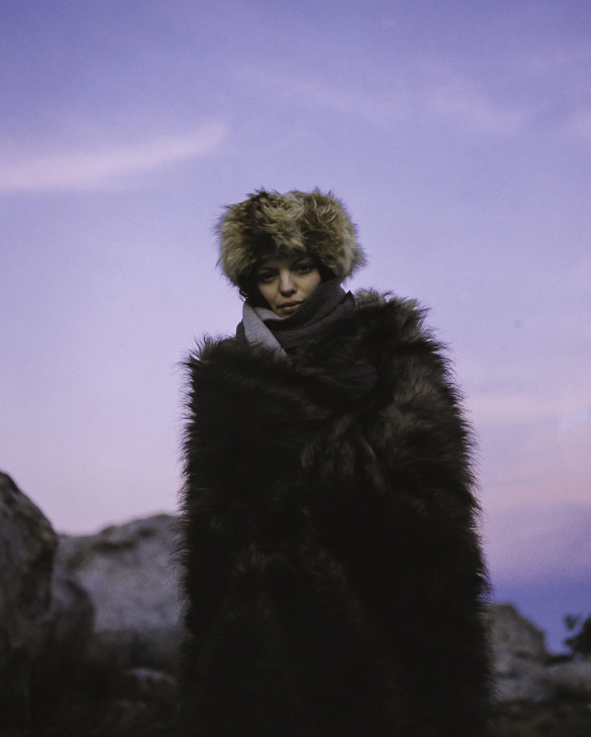 A person dressed warmly in a fur hat and coat, standing outdoors under a clear, purple sky.