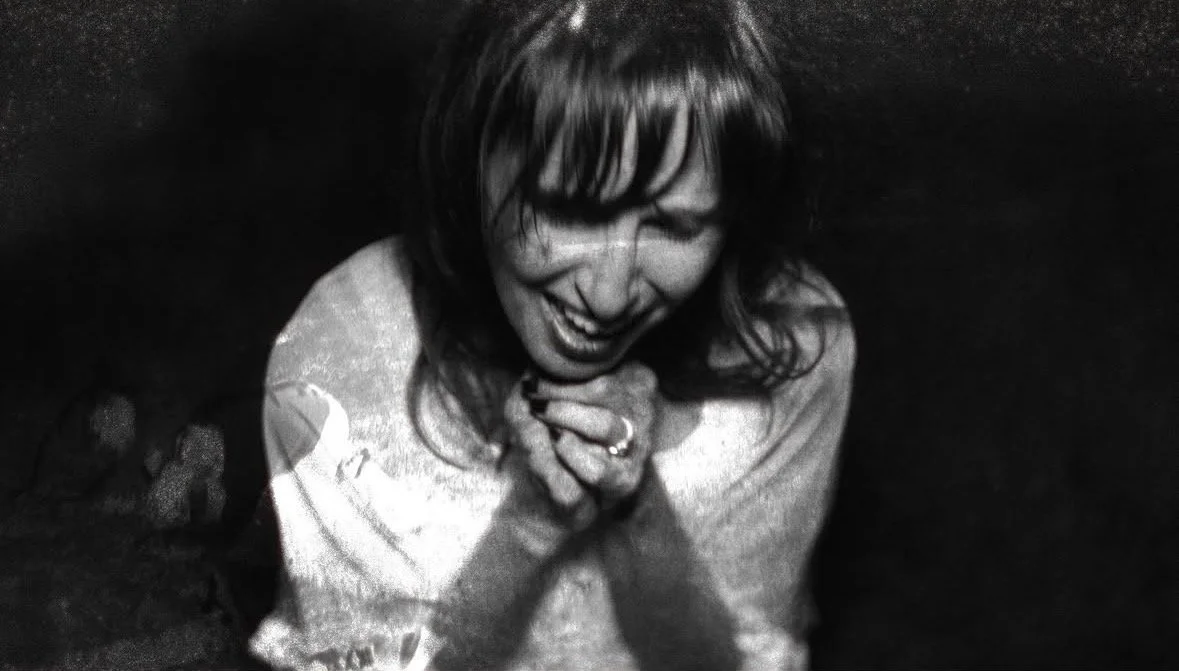 A woman laughing and smiling with her hands clasped together near her face in a dark setting.