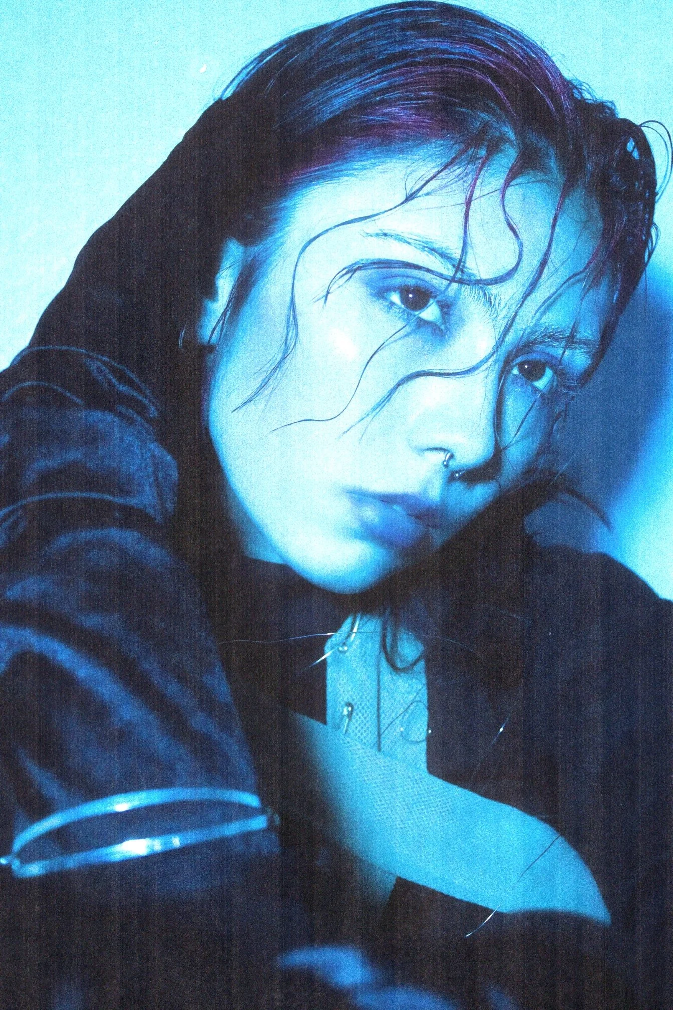 A close-up portrait of a woman with wet, dark hair and pierced nose, wearing a striped shirt and a choker necklace, with a serious expression.