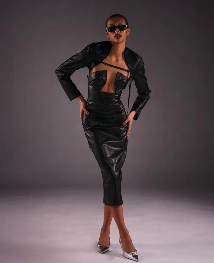 Fashion model wearing a black leather dress with cutouts, a matching cropped jacket, sunglasses, and silver high heels, posing with hands on hips against a gray background.