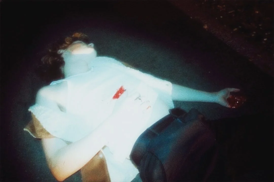 A person lying on the ground in a dark environment, wearing a white shirt with red marks, black pants, and glasses, with one arm extended.