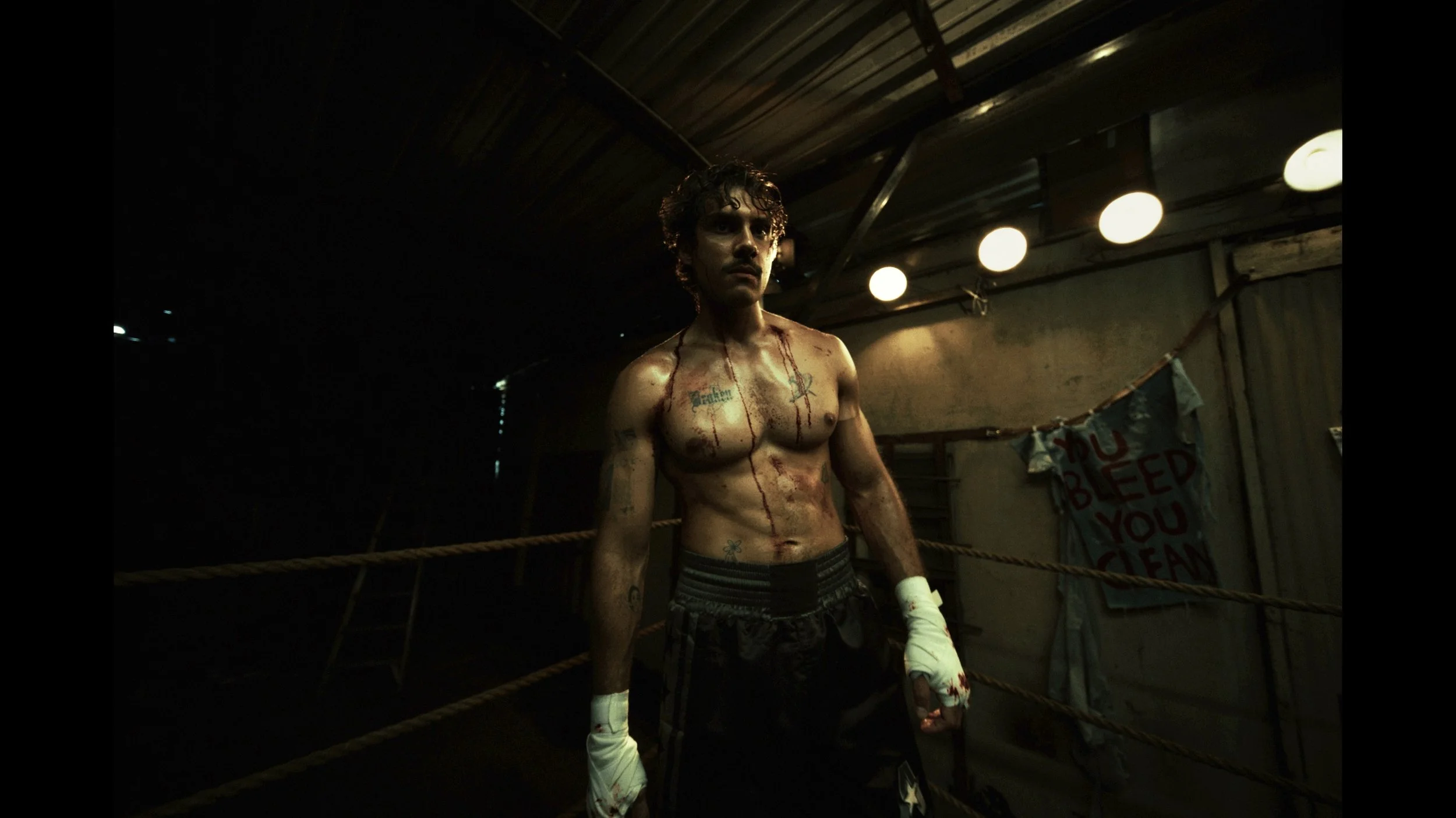A shirtless man with curly hair and tattoos on his chest and arms, standing in a dimly lit boxing ring with blood on his body and wrapped hands, in a gritty environment with hanging lights and a banner in the background.
