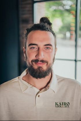A man with a bun hairstyle and beard, wearing a beige shirt with a logo reading 'KAIRO LOCK & KEY', sitting indoors near a window with a view of trees outside.