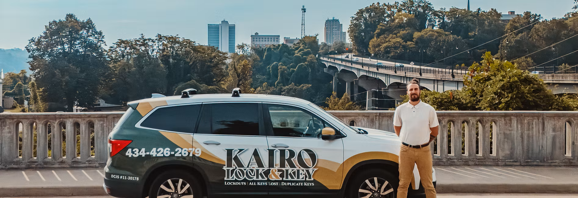 A man standing next to a locksmith service vehicle parked on the street with trees, a bridge, and city buildings in the background.