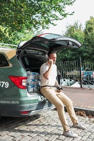 A locksmith from Kairo Lock & Key sitting on the open trunk of a green SUV while speaking on the phone, with tools and water bottles visible, parked on a city street during a mobile service call.