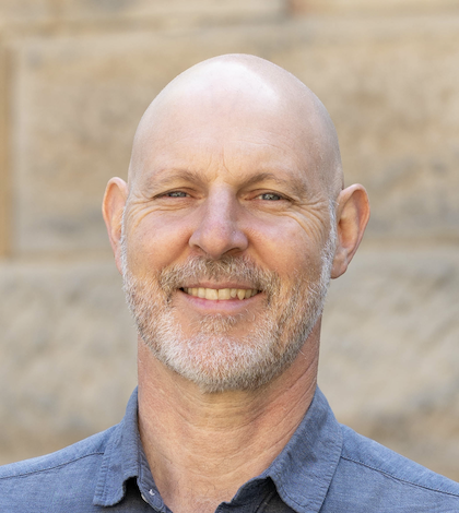 Portrait of a middle-aged bald man with a beard smiling outdoors in front of a stone wall.