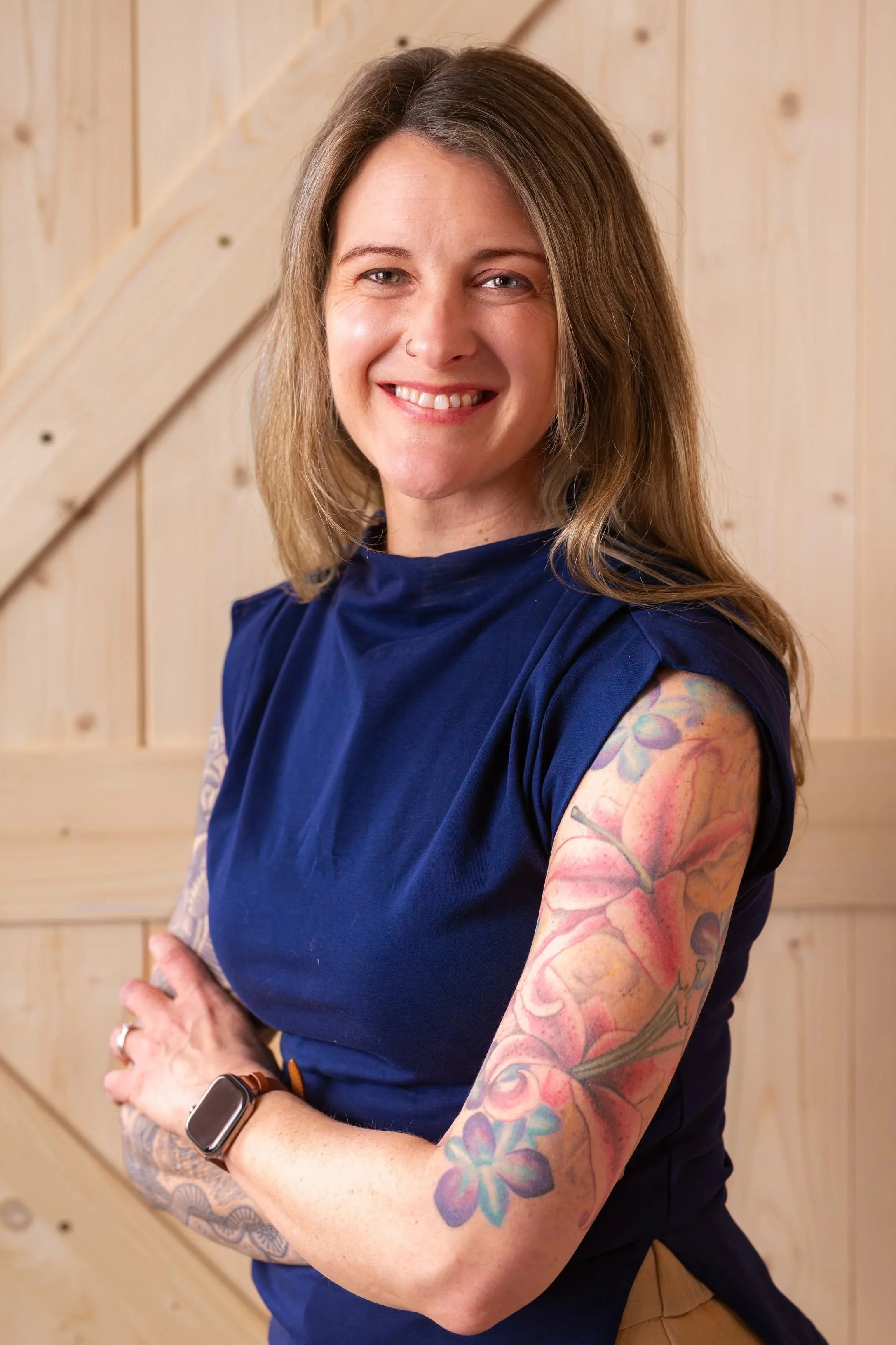 A smiling woman with long light brown hair, wearing a navy blue top, showing colorful tattoos on her left arm, standing in front of a light wooden backdrop.