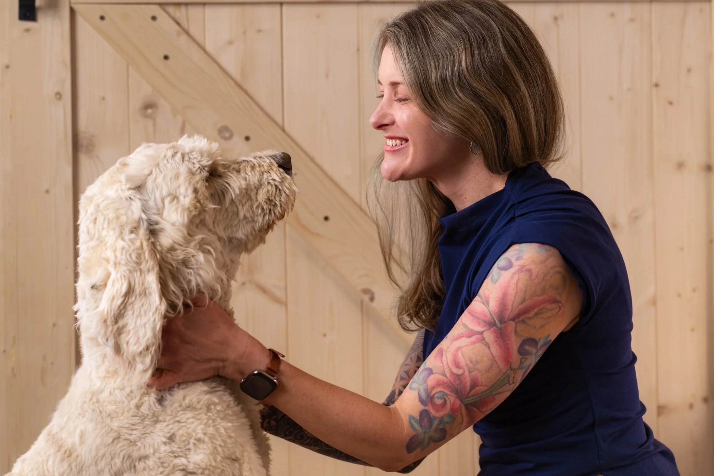 A woman with tattoos on her arm and shoulder smiling as she holds a fluffy, light-colored puppy near her face. They are indoors with wooden panel walls in the background.