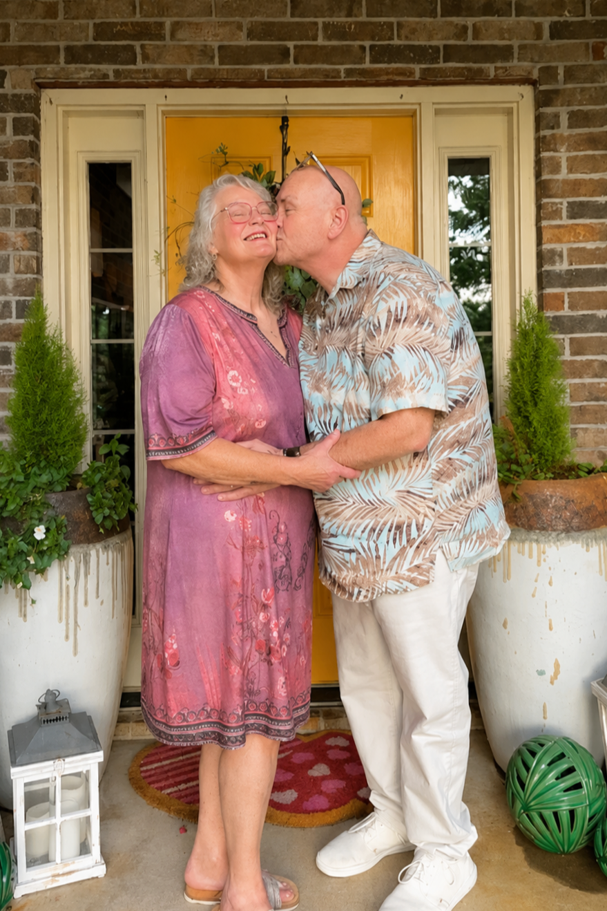A man and woman sharing a kiss and smiling on a porch in front of a yellow door, surrounded by potted plants and decorative items.