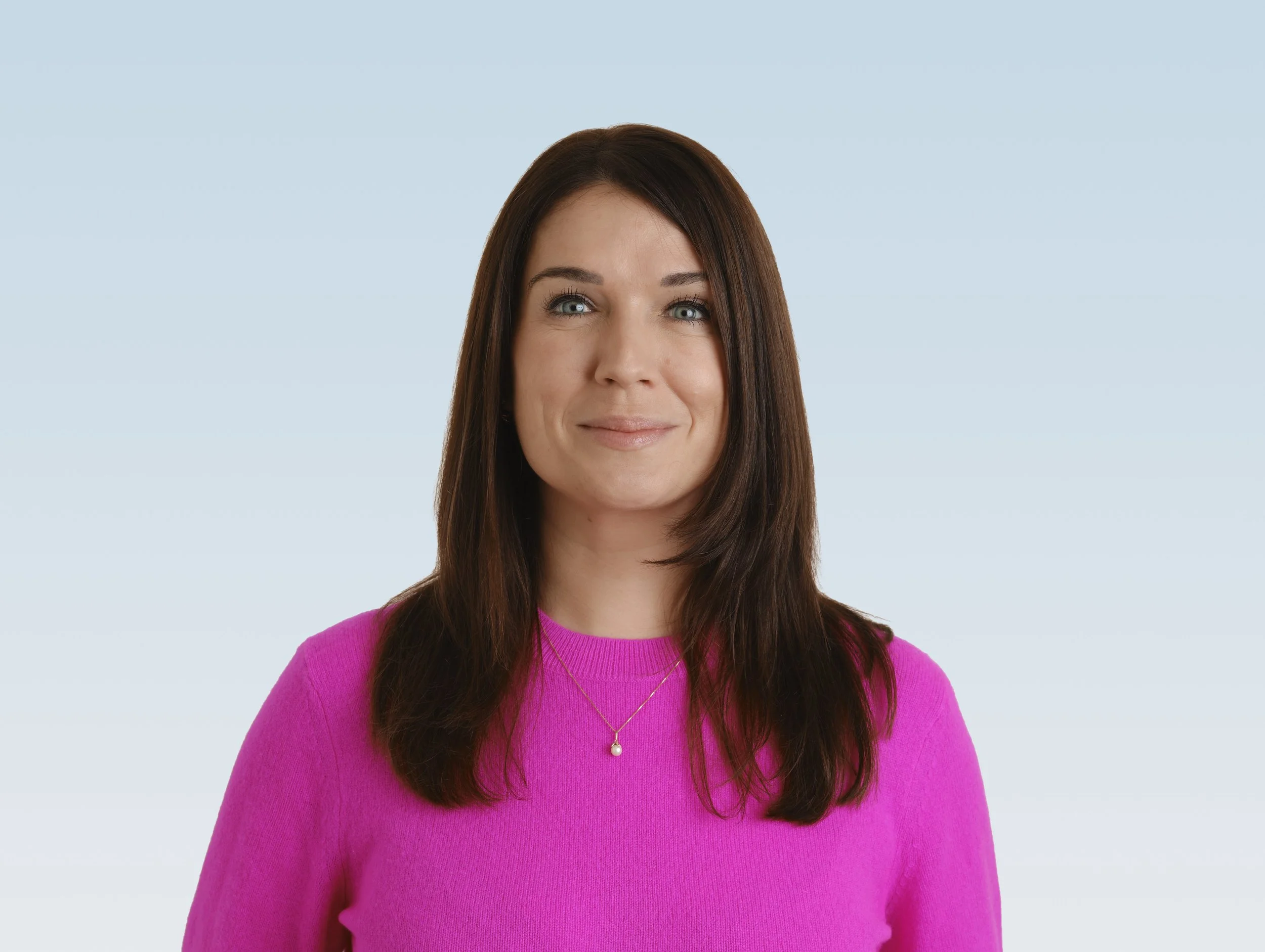 A woman with long dark hair wearing a bright pink sweater and a pearl necklace, standing against a light blue background.