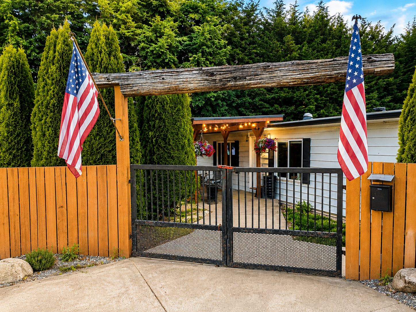 Front yard of a house decorated for the 4th of July, featuring two American flags, a black metal gate, a wooden fence, and a small patio with hanging flower baskets and string lights.