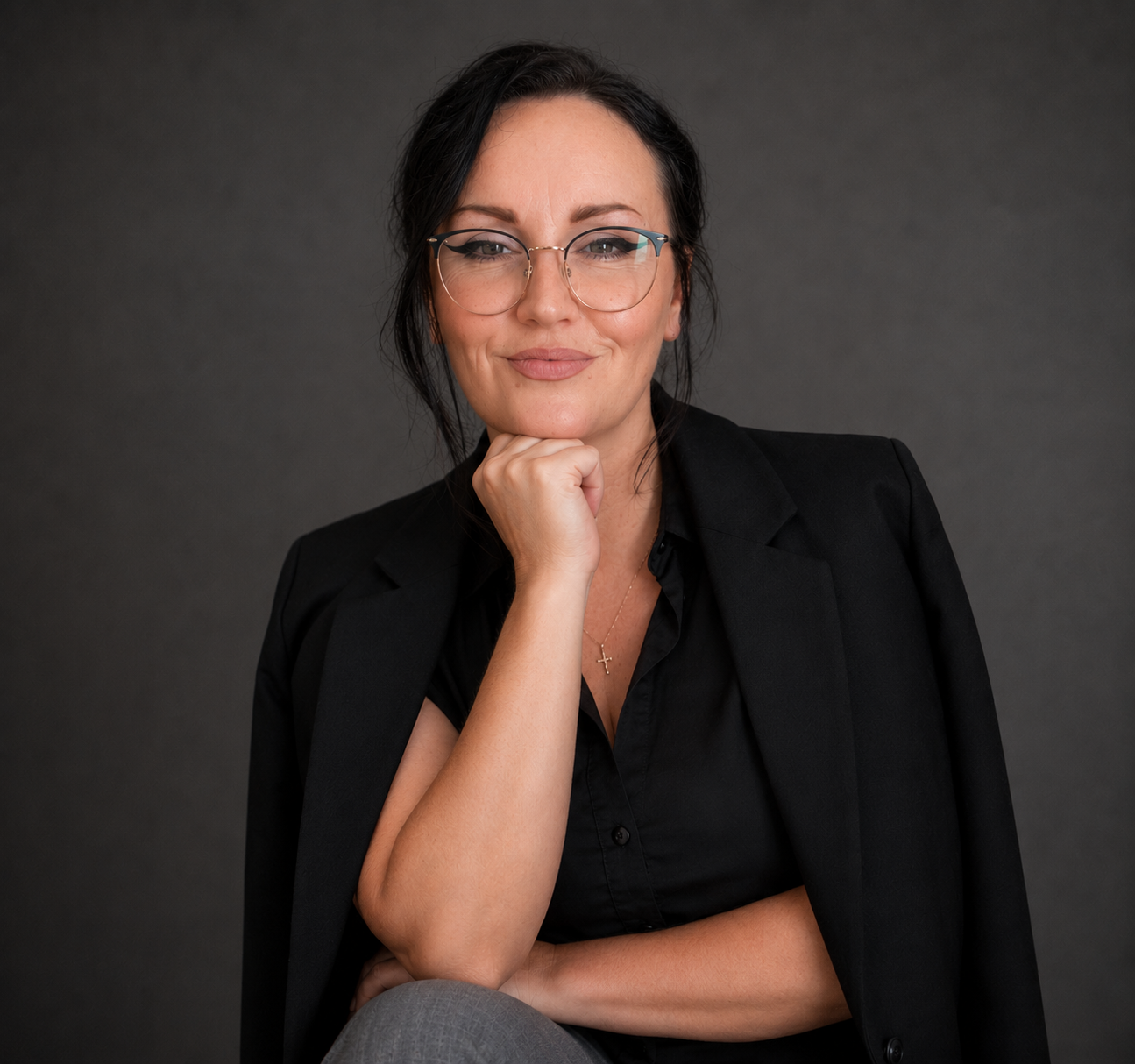 A woman with dark hair and glasses, dressed in a black blazer and blouse, sitting against a plain gray background, with her chin resting on her hand and a confident expression.